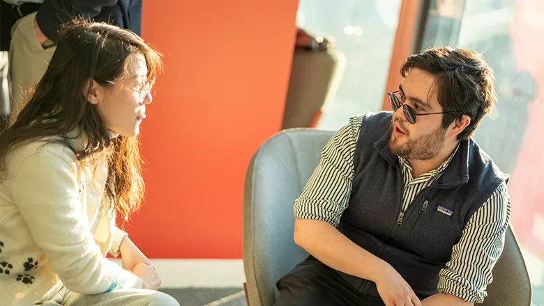 Francis wearing a pair of sunglasses, a lined shirt, sitting as he talks with a person with long brown hair. 