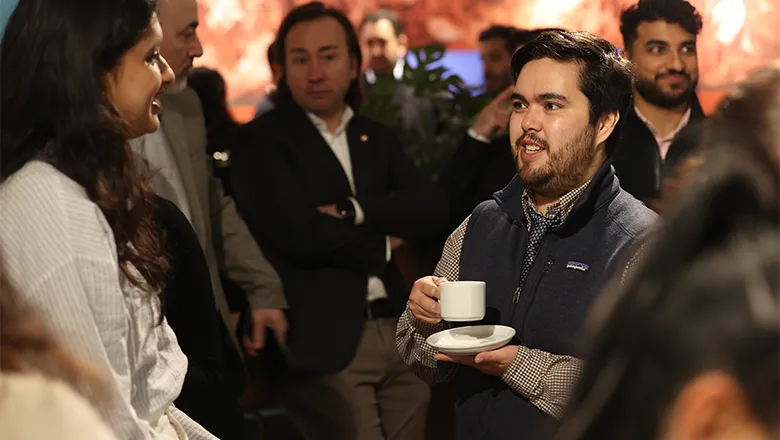 Francis holding a cup of hot drink, while speaking to a person with long dark hair. The background shows a diverse group of people in a networking event. 