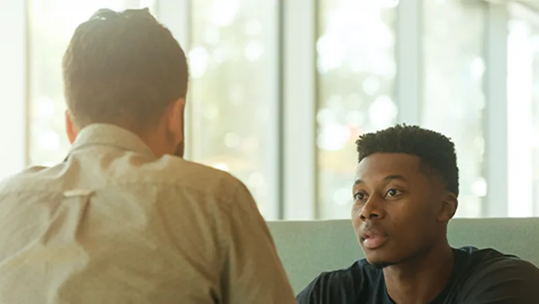 A mentor sits with his back to the camera, facing a young Black man who is the mentee in this relationship. They are sat at a table talking in a well-lit room by some windows.