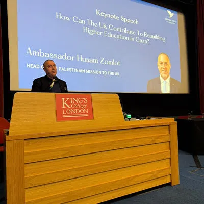 A man in a dark suit stands at a podium inside a lecture theatre at King’s College London. On a large screen behind him is an image with a caption that identifies the speaker as Dr Husam Zomlot, Palestinian Ambassador to the UK 