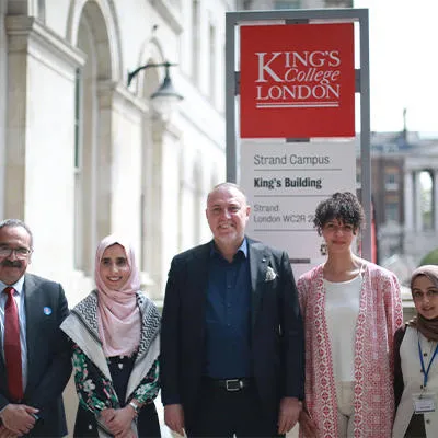 A group of five people in smart, formal clothes stood outside a building on The Strand campus of King’s College London. Two of these are men in suits and three of them are women. 