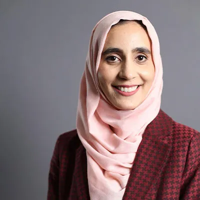 A young woman wearing a red coat and a pink headscarf smiles at the camera 