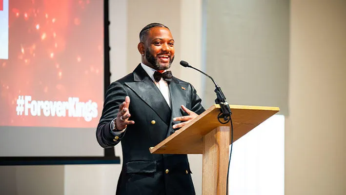 A man wearing a black tuxedo stands at a lectern and warmly talks to an audience. Behind him is a screen with the words ‘King’s Distinguished Alumni Awards 2025’