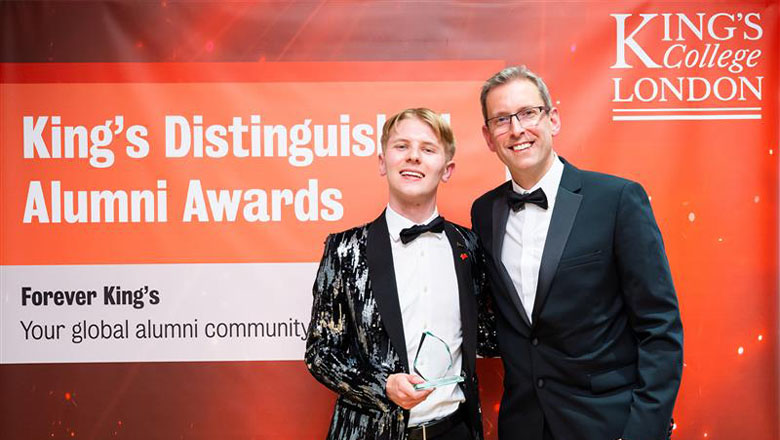 A young man in a spectacular silver and black tuxedo, white shirt and bow tie holds an award in his hands. Standing next to him is a middle-aged man in a tuxedo. Behind them is a backdrop that read King’s Distinguished Alumni Awards.