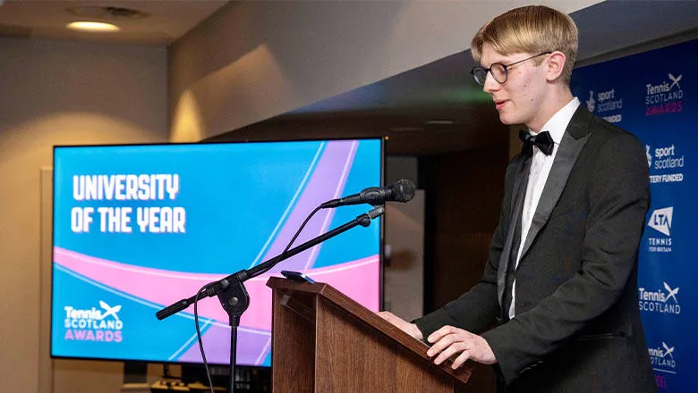 A young man in a tuxedo, white shirt and bow tie stands on stage at a lectern giving a speech to an audience at an awards ceremony. A screen behind him has the words ‘Tennis Scotland Awards’ and ‘University of the Year’