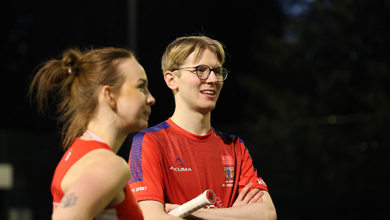A young man with glasses and short hair stands holding a tennis racket under his arm. Next to him is a young woman with long dark hair. Both wear red sports tops with the King’s College London logo on their chests.