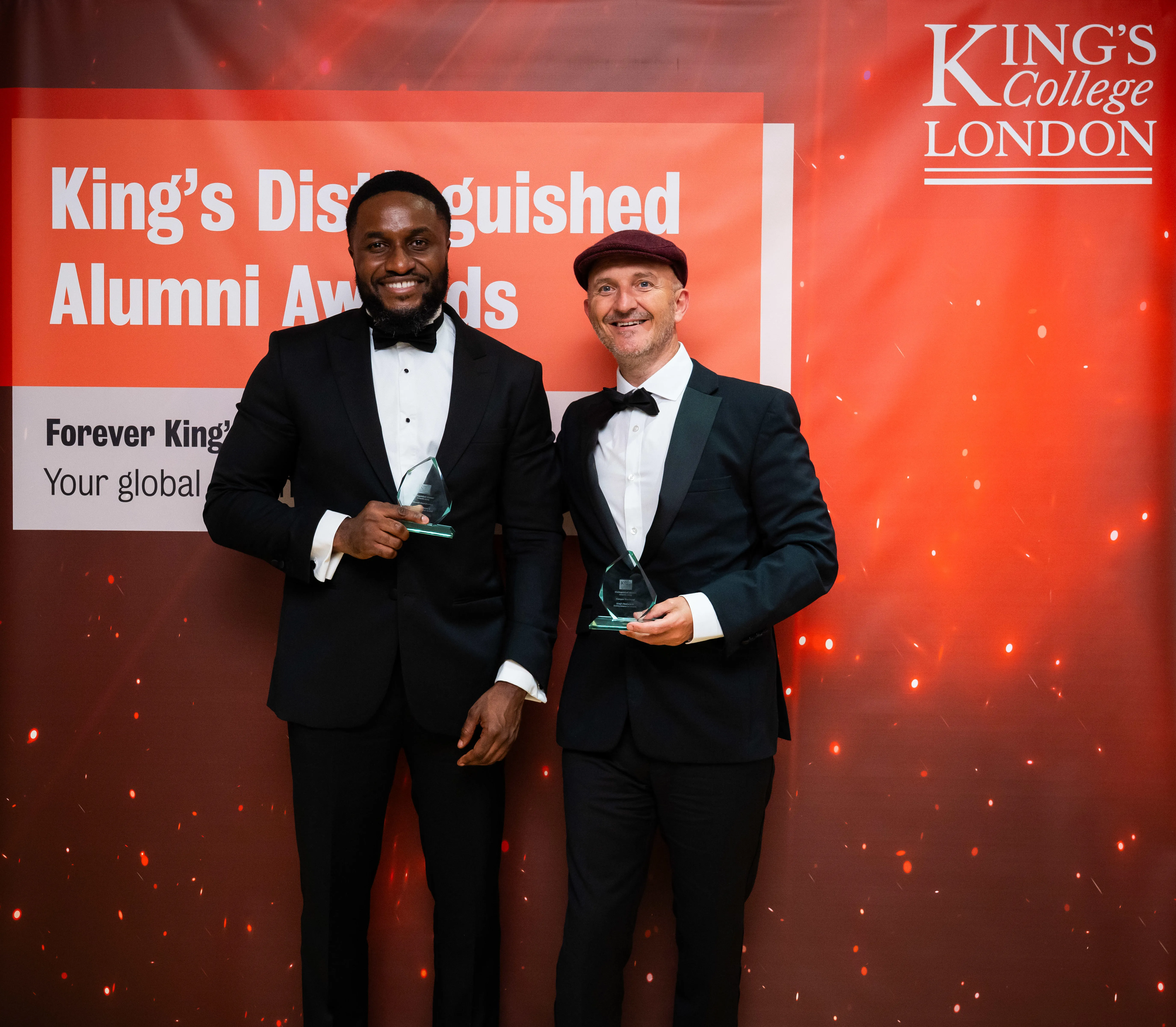 Two men wearing black tuxedos stand in front of a screen with the words ‘King’s Distinguished Alumni Awards 2025’. They both smile and hold awards in their hands. The man on the left wears a flat cap.