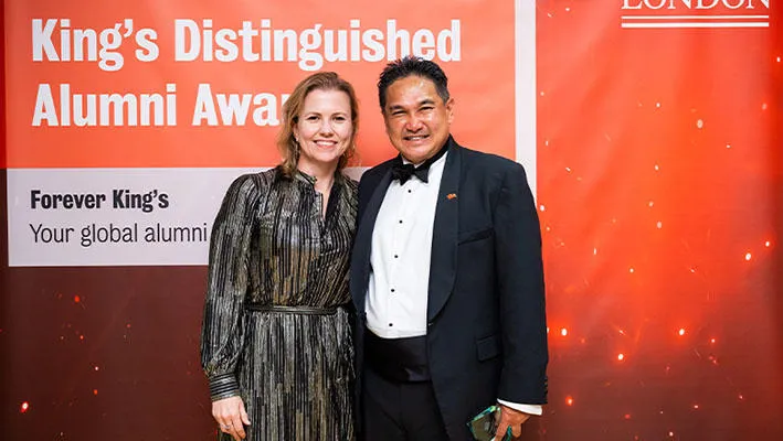 A young woman in a stylish silver and black dress stands next to a man with short, dark hair wearing a black tuxedo. They stand in front of a screen with the words ‘King’s Distinguished Alumni Awards 2025’. They both smile. He holds an award in his hands.