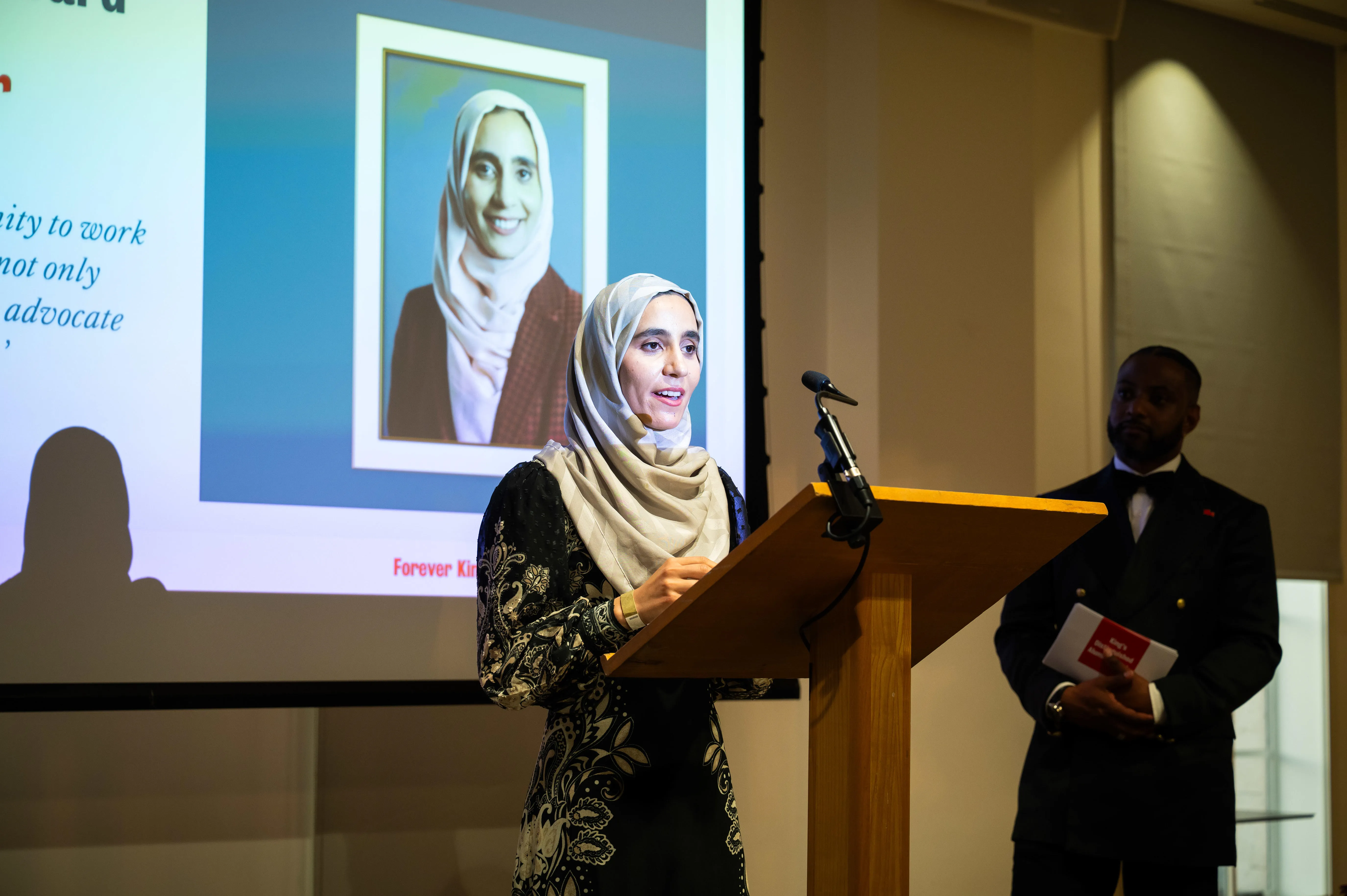 A young woman wearing a patterned black dress and a headscarf stands on stage at a lectern in front of a screen. She addresses an audience.