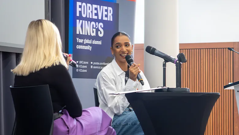 Laviai Nielsen sits in the Great Hall at King's delivering a talk. She wears an oversized white shirt and jeans and sits in front of a banner reading 'Forever King's'.