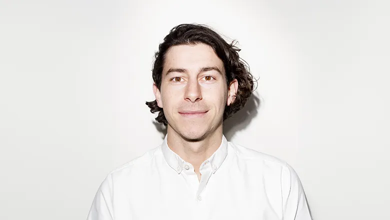 A young man with shoulder-length dark hair. He is smiling at the camera and he wears a white shirt.