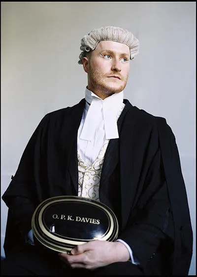 A head shot of Oscar in a barrister's gown and wig, holding a name plate titled 'O. P. K. Davies'.