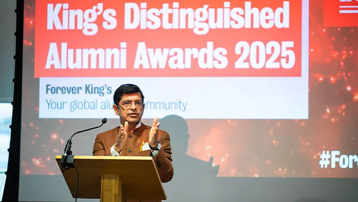 A man with short dark hair and a brown jacket stands at a lectern. Behind him is a screen with the words ‘King's Distinguished Alumni Awards 2025’