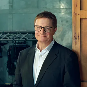 Alex Beard, wearing a black suit and large glasses, smiling slightly. He has a neatly styled hair and is posed against a blurred green backdrop.