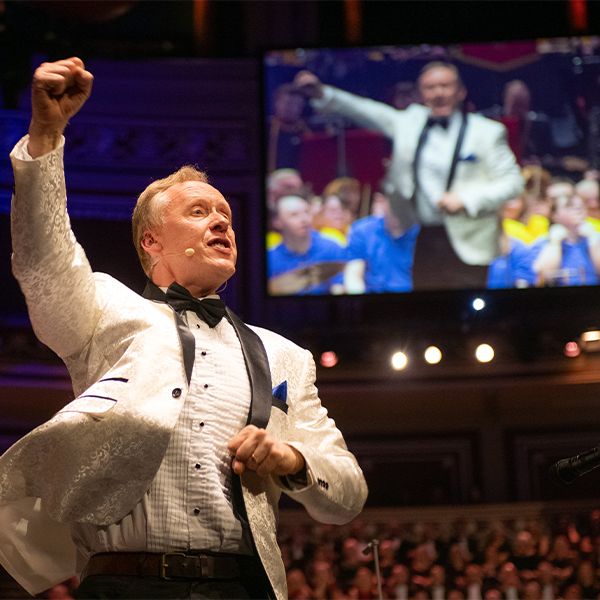 David Stanley BEM wearing a white patterned suit jacket, black bow tie, and microphone headset raises his fist enthusiastically while standing on stage in front of an audience. A large screen behind him shows his image as he performs in a brightly lit concert hall.