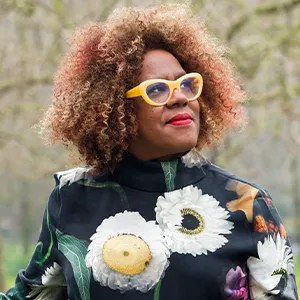Errollyn Wallen, a person with curly hair wearing bright yellow glasses and a black floral top with large white and yellow flowers.
