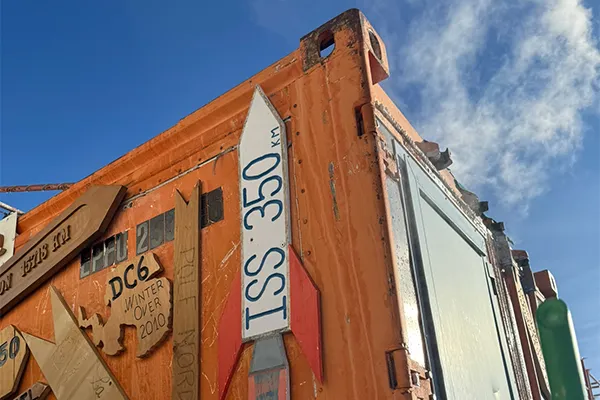  The image features a weathered orange wall with various wooden signs and pieces attached to it. Among the items is a rocket-shaped sign labelled ‘ISS 350’. The background shows a blue sky.