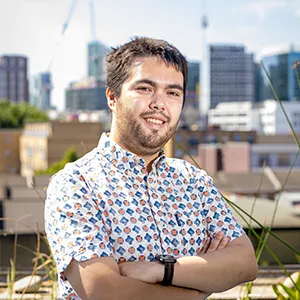 Francis Jones with short, tousled hair and a beard, smiling while standing outdoors with the city skyline visible in the background. He is wearing a patterned shirt and has his arms crossed.