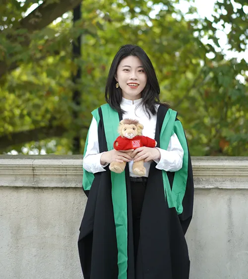 An image of King's College graduate Chang Xu. She is wearing her graduation robe, and holding a toy version of Reggie, the King's college London mascot.
