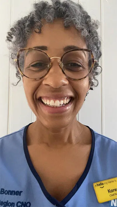 A woman with grey hair and glasses wearing a blue nursing uniform. This uniform has the words ‘Karen Bonner London Region CNO’ on the right side 