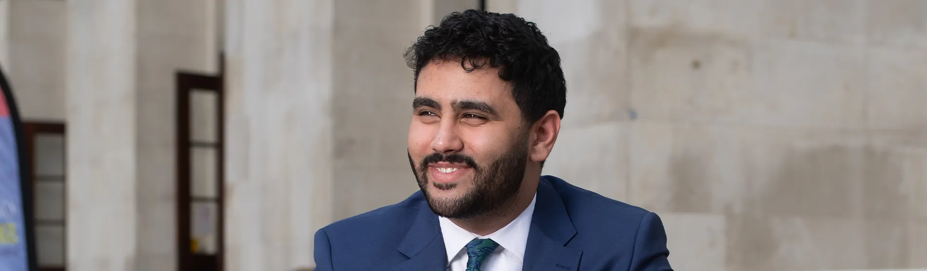 Mohamed Elhag is smiling to the left of the camera wearing a crisp blue suit.