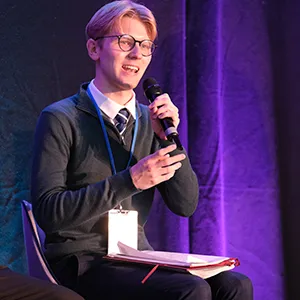 Jonathon Dawes with short brown hair and round glasses, smiling while holding a microphone. They are wearing a dark sweater over a white shirt and are seated on stage with a purple backdrop.