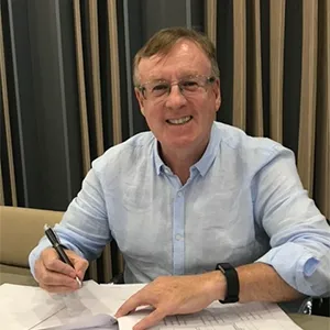 Joseph Healy with short, wavy hair and glasses, smiling brightly while seated at a table. He is wearing a light blue shirt and holding a pen while writing on a piece of paper.