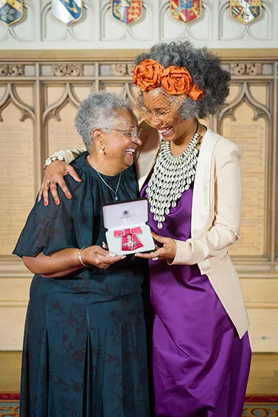 On the right is a woman wearing an elegant purple dress with a white jacket. On the left is an older woman wearing a dark turquoise dress. The two hug each other affectionately. Between them, they jointly hold a medal housed in a small black box. 