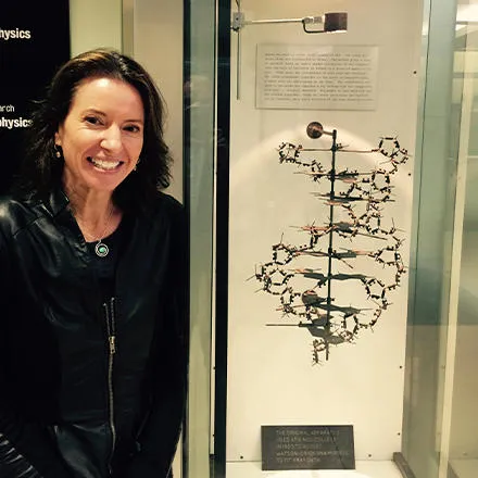 A woman with long brown hair and wearing a black leather jacket stands next to a display cabinet. This houses a scientific model showing the structure of DNA. 