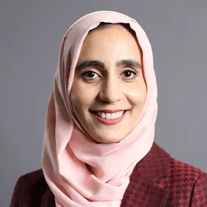 Dr Khaleda Zaheer, wearing a pink hijab and a plaid blazer, smiling warmly and standing against a grey backdrop.