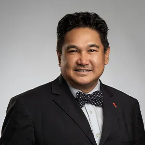 Kelvin Miranda smiling in a formal black suit and a bow tie. He has short, dark hair and is posed against a grey backdrop.