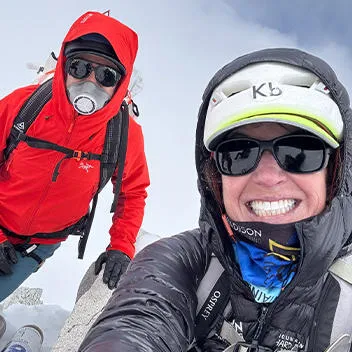 Two climbers scale the snow-covered peak of Mount Makalu in the Himalayas. One wears a bright red heavily insulated jacket and the other wears a heavily insulated black jacket