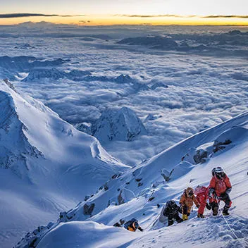 A group of mountaineers make their way up a snow-covered peak. In the background is a vast expanse of snow-covered mountains. 