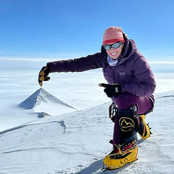 A woman in climbing boots and wearing a heavily insulated purple mountaineering jacket and trousers, goggles and a beanie hat makes a playful pose near the top of a snow-covered peak ntaineering