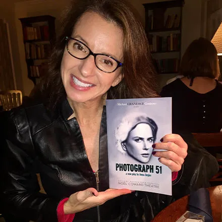 A woman with long brown hair and wearing glasses, a black leather jacket and leopard print trousers holds up a play programme. The cover reads ‘Photograph 51’. 