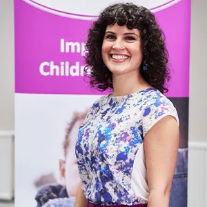 A white woman with frizzy black hair, wearing a patterned blue and white blouse, smiles.