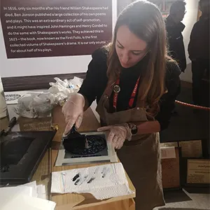 A brunette woman wearing a red lanyard, black top and brown apron, performs crafts.