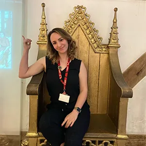 A brunette woman wearing a black outfit sits in a golden throne, pointing up at the air.