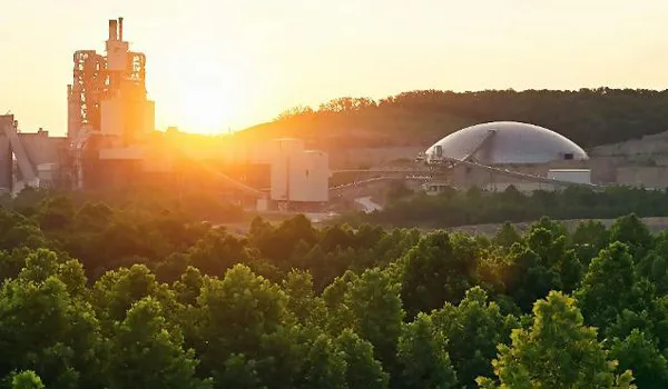 The sun sets behind an industrial site located amongst green trees.
