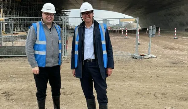 Two white men in hard hats and blue high visibility jackets smile at the camera.