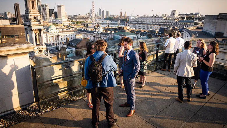 People mingle on the rooftop of Bush House, with the London skyline in the background.