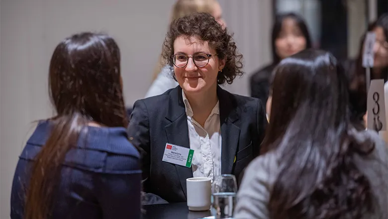 A woman with short brown hair and glasses, wearing a smart suit, speaks to two students at a networking event.