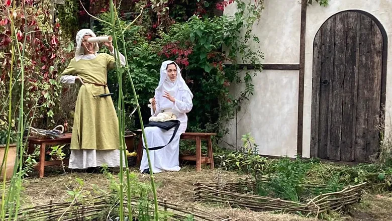 Two actresses in medieval dress, one a yellowy green and the other in a white one, sit near a wooden door.