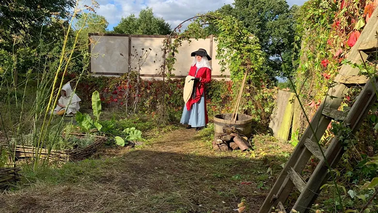 A woman wearing a medieval red dress stands in a garden.