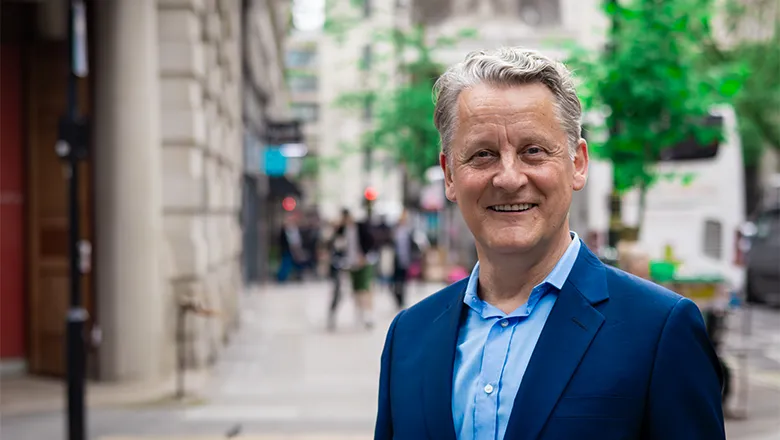 A man stands on a busy London street, smiling and wearing a stylish navy suit jacket over a lighter blue shirt.