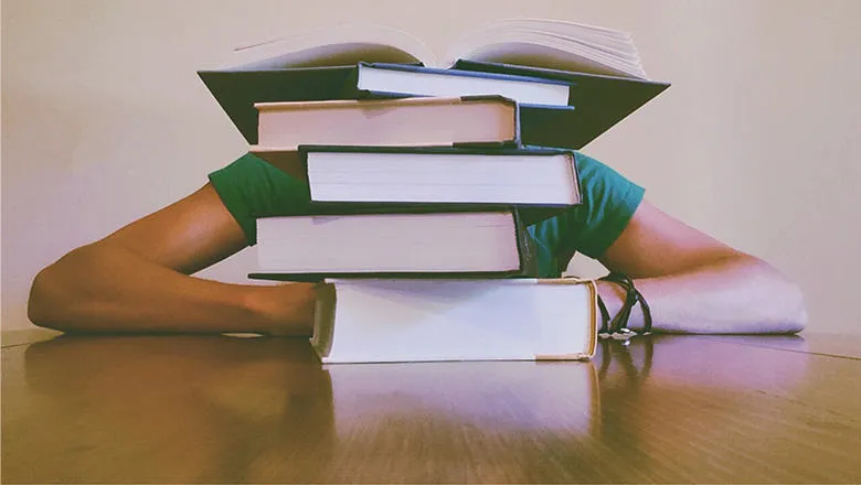 A pile of books on a wooden tabletop with a person hidden behind.