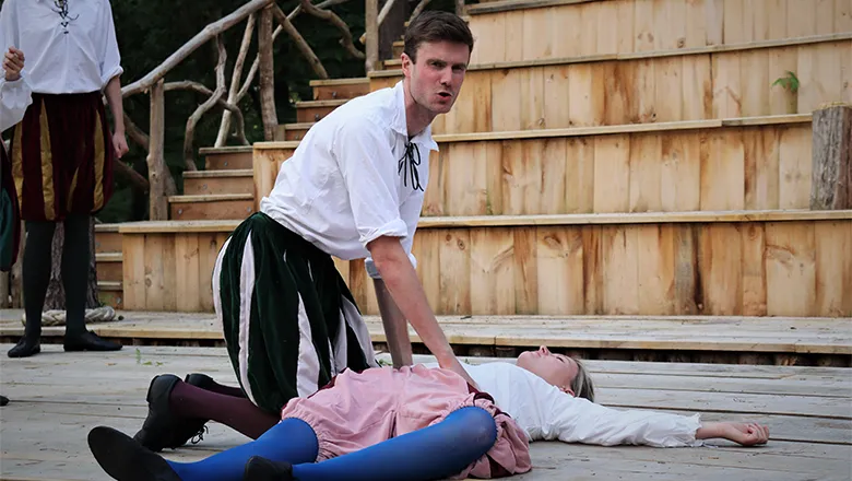 A young man dressed in Elizabethan costume is on stage, kneeling over the body of a young woman also in Elizabethan costume