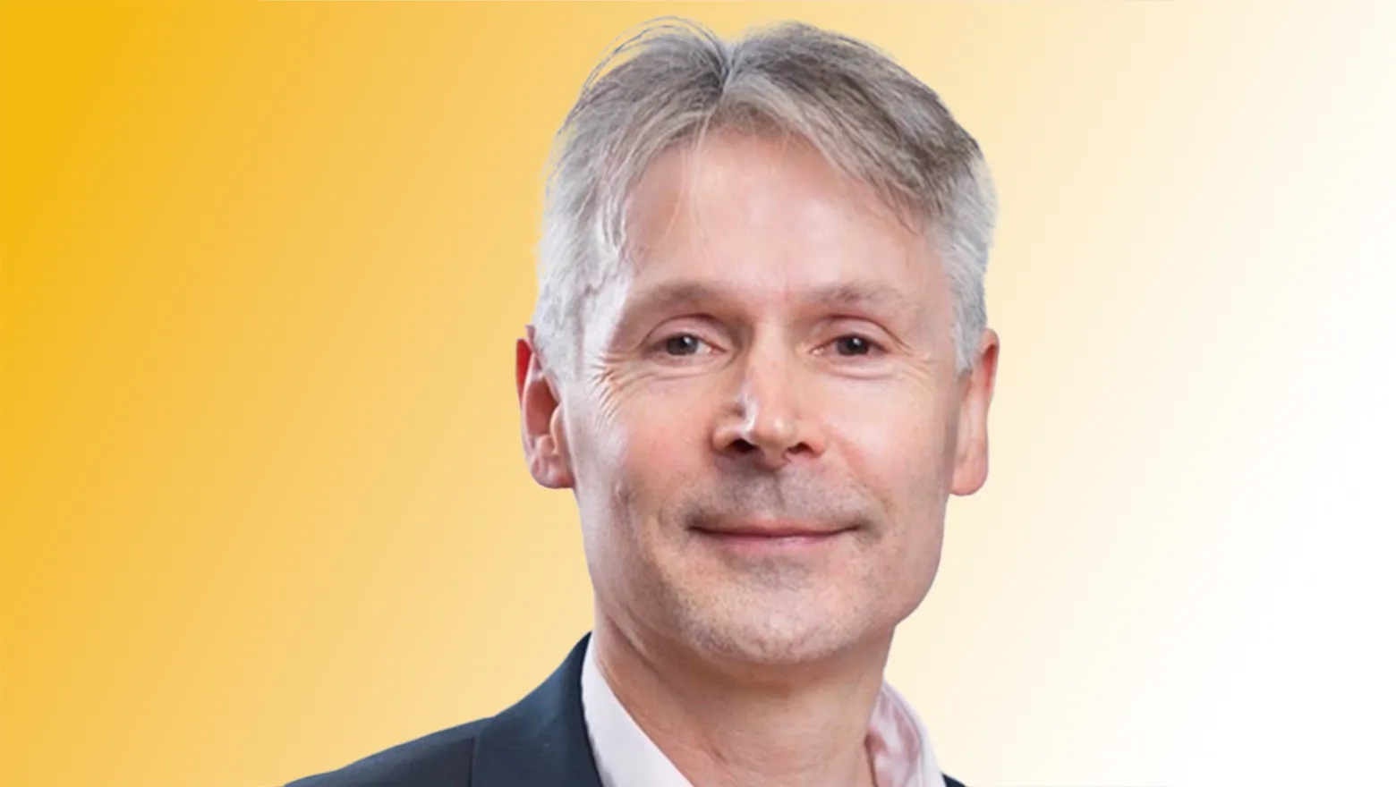 An image of Neil Braithwaite. He is wearing a white shirt and a dark suit, and standing against a yellow background.