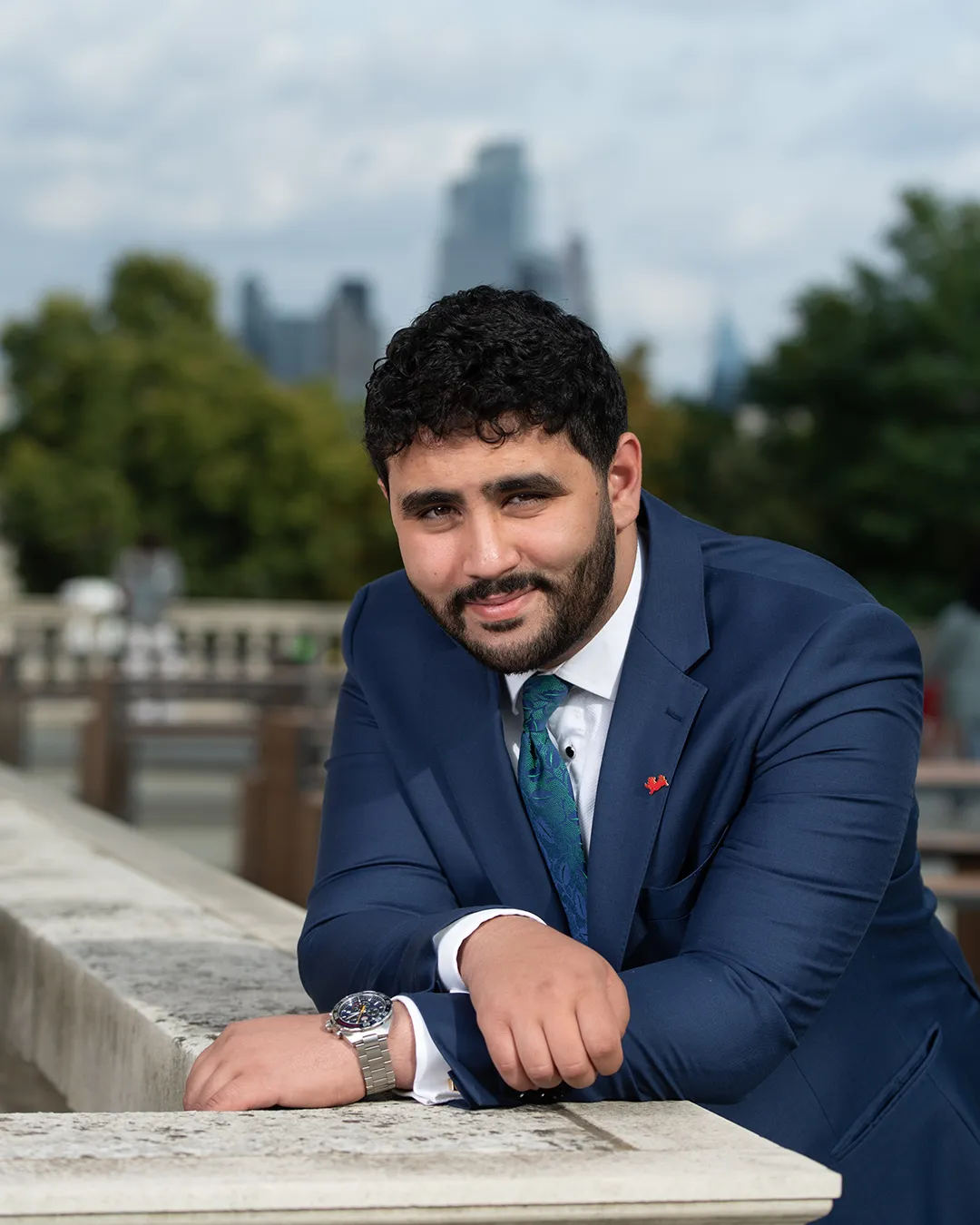 Mohamed Elhag stands and rests his arms on a white marble plinth with the London skyline behind him. He is dressed in a blue navy suit.