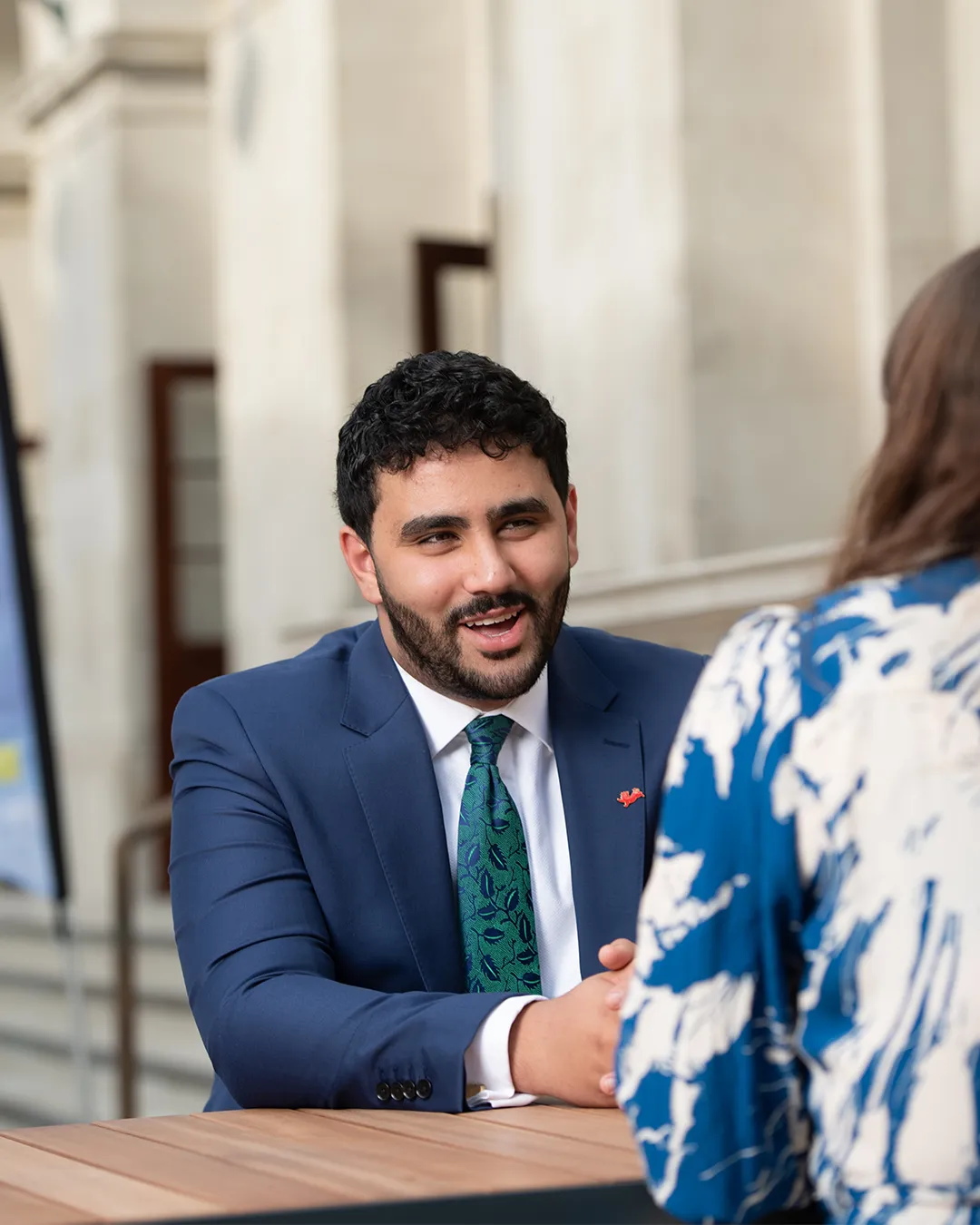 Mohamed Elhag is sat at a bench dressed in a blue suit talking to someone wearing a splotchy blue and white dress who has their back turned to the camera.